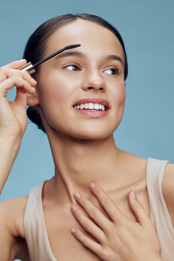 Young woman with natural makeup applying mascara, showcasing beauty routines and self care while expressing joy against a soft blue background